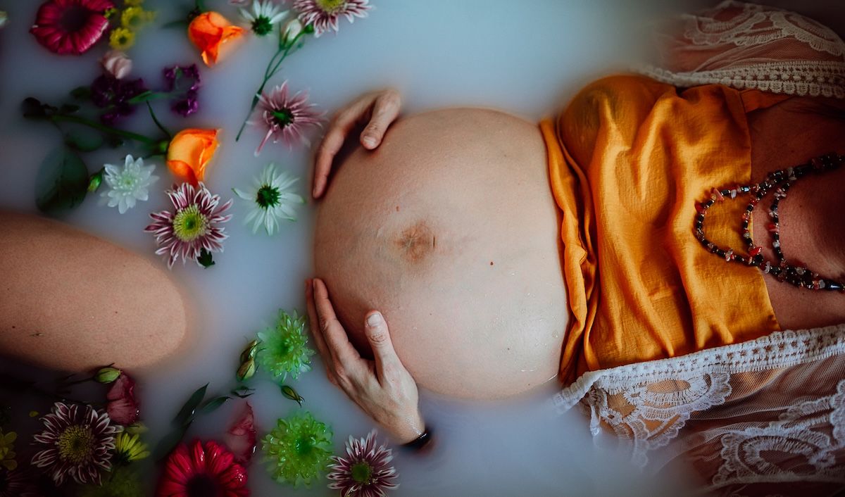 a closeup of a pregnant woman's bump while she lays in a milky bath with flowers surrounding her. 