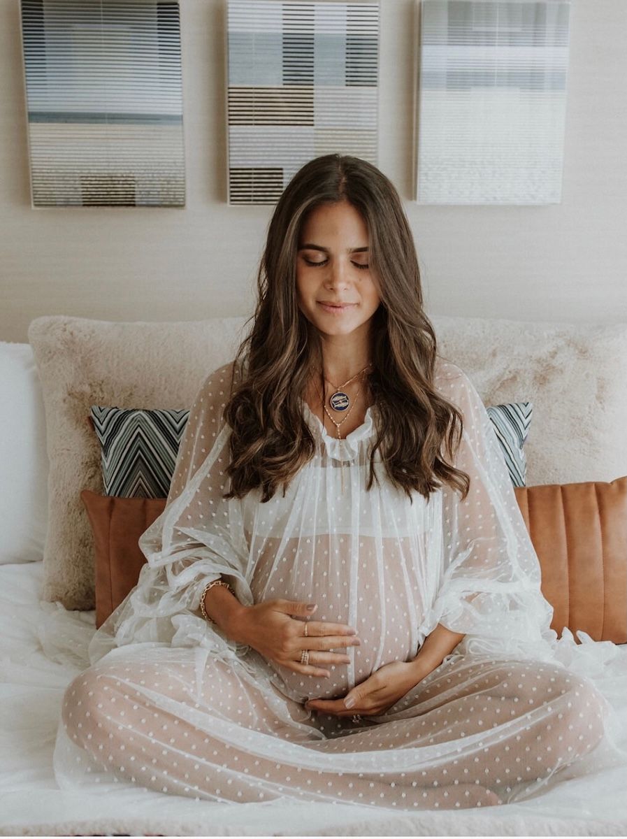 a home maternity shoot featuring a woman sat cross legged on her bed in a lacy dress holding her bump