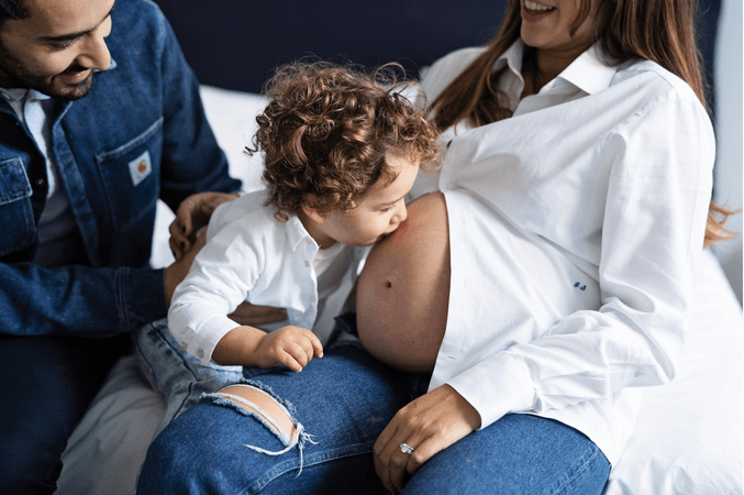 Maternity shoot featuring a couple and child sitting on a bed, the young boy kisses his pregnant mum's stomach