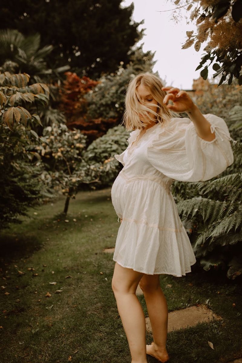 a candid shot of a pregnant woman standing in a garden wearing a white dress, hair in front of her face