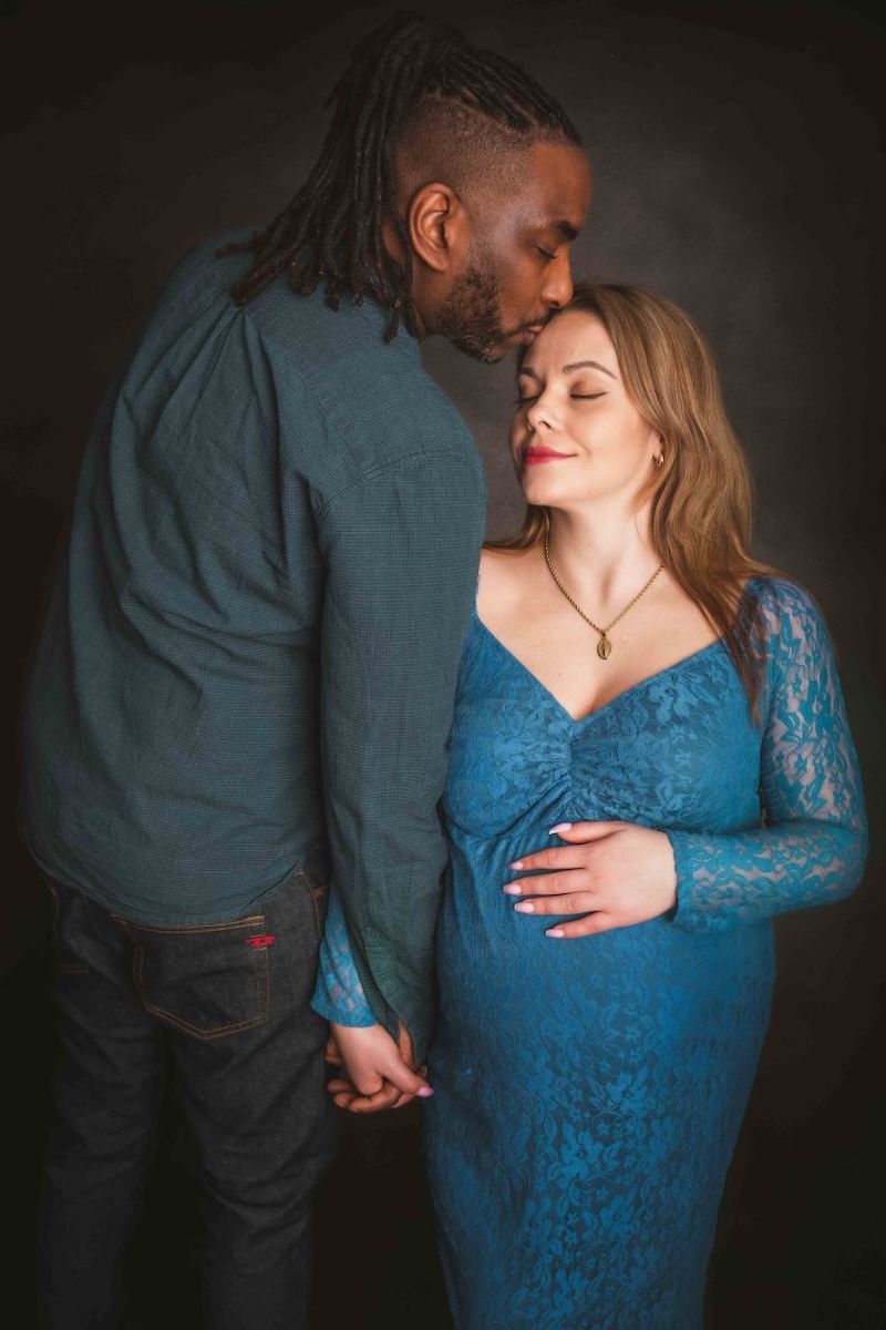 A studio maternity portrait of a couple, the man kisses the top of the pregnant woman's head while she holds her bump