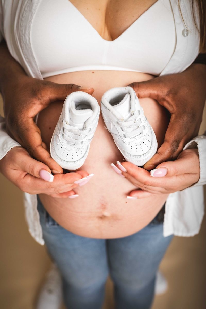 close up of pregnant woman's stomach while her and her partner hold up a pair of white baby sneakers in front of her