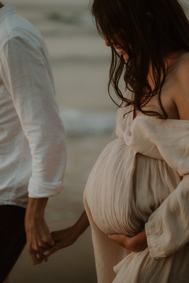 close up  of couple holding hands on the beach while the woman holds her baby bump