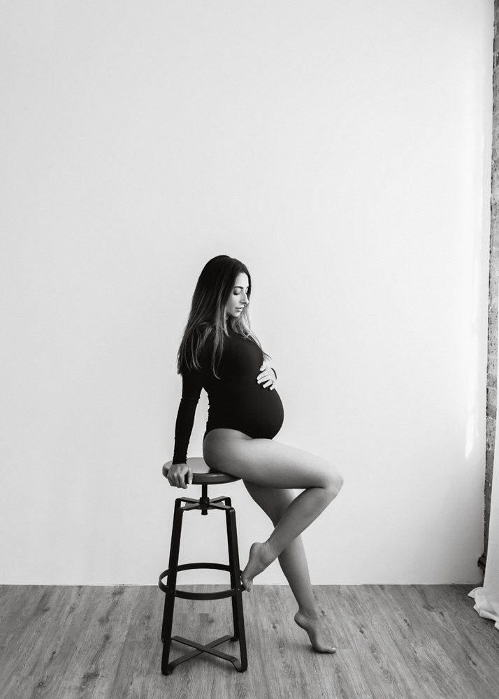 black and white studio image of a pregnant woman in a black body suit, posing on a stool