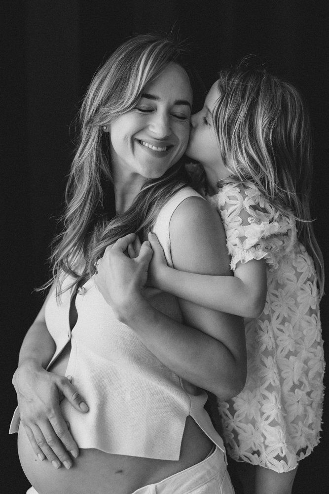 black and white image of a young girl kissing the cheek of her smiling pregnant mum