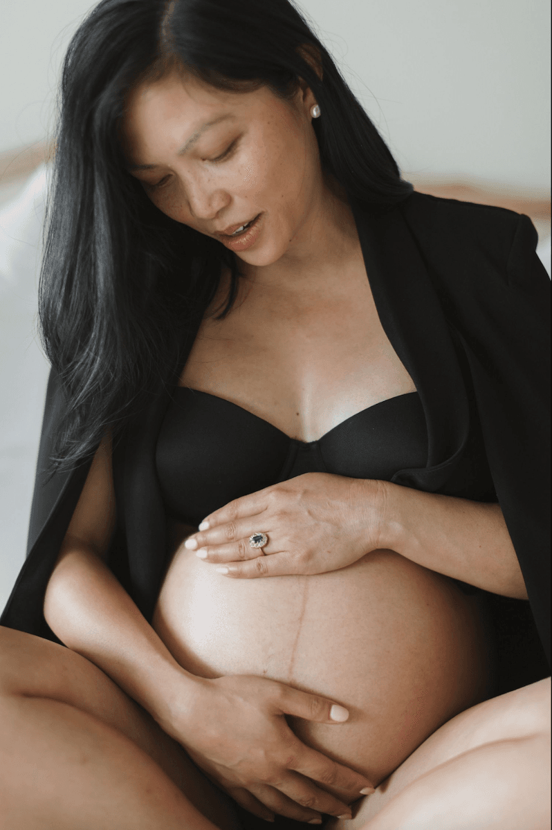 Woman dressed elegantly in black, sitting cross legged on her bed holding her baby bump