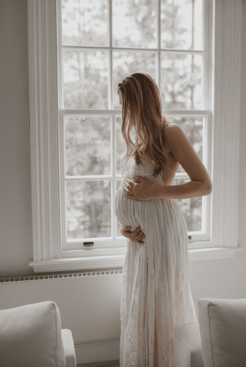 woman standing next to a window in her house in a long lacy dress, holding her baby bump