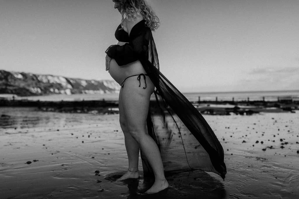 black and white image of a pregnant woman walking barefoot on wet sand wearing a bikini 
