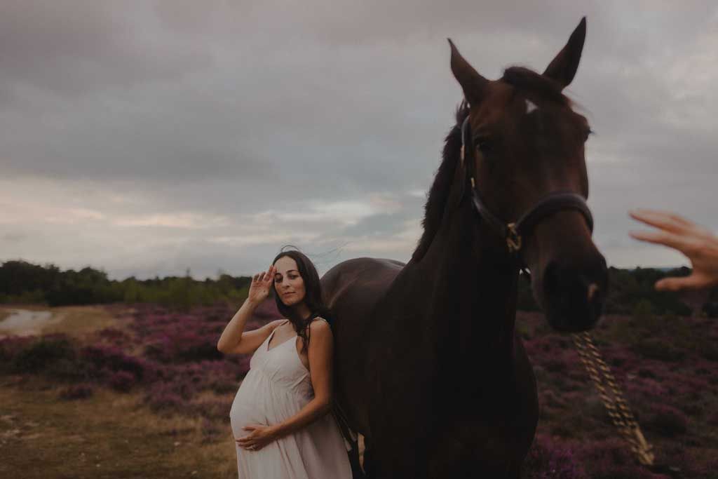 pregnant woman standing next to a horse in the countryside on an overcast day