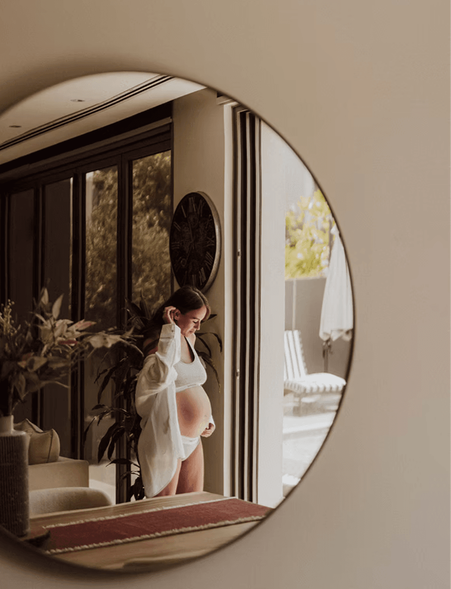 mirror reflection of a pregnant woman standing in her home in underwear and a white shirt
